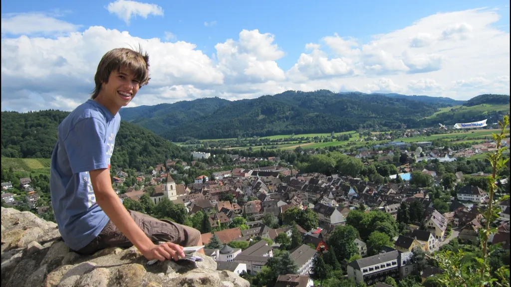 Jonas sitzt freundlich lächelnd hoch oben auf der Brüstung einer Burg über einen kleinen Stadt in Baden